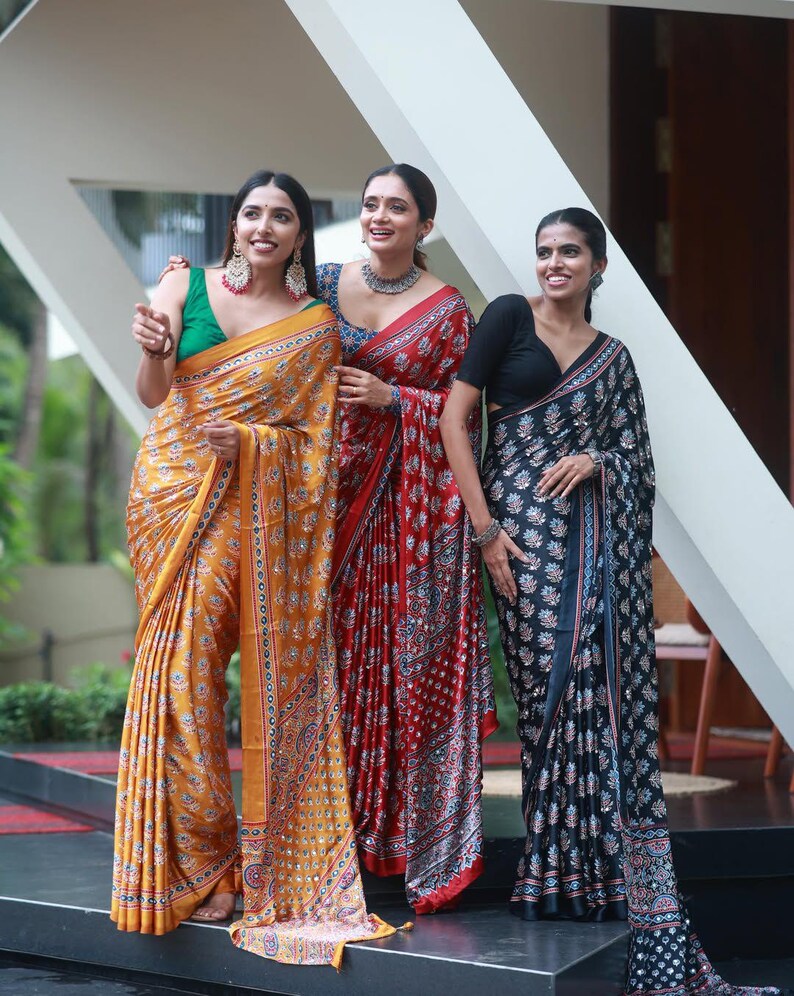 May include: Three sarees featuring intricate floral patterns and reflective mirror work. One saree is mustard yellow with a green blouse, one is deep red with a blue blouse, and one is black with a black blouse, all draped in traditional style.