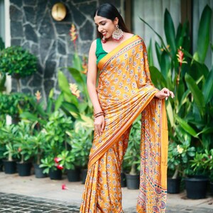 May include: A mustard yellow saree featuring a repeating floral pattern in blue, white, and red, paired with a solid green sleeveless blouse. The saree has a decorative border with intricate geometric and floral motifs in matching colors.
