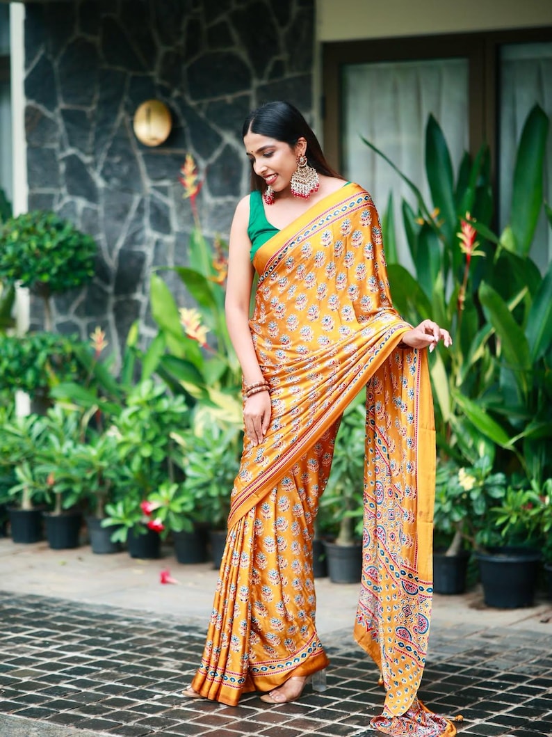 May include: A mustard yellow saree featuring a repeating floral pattern in blue, white, and red, paired with a solid green sleeveless blouse. The saree has a decorative border with intricate geometric and floral motifs in matching colors.