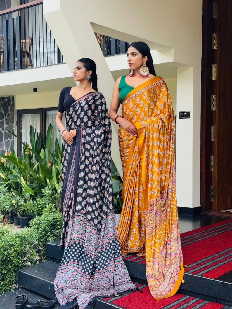 May include: Two sarees, one black with a white and pink floral pattern and a solid black blouse, and one mustard yellow with a white floral pattern and a solid green blouse. Both garments feature intricate borders and draped fabric silhouettes.