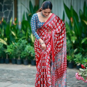 May include: A red saree featuring a repeating white floral block print pattern with a blue and white geometric patterned blouse. The saree has a wide border with intricate blue, white, and red circular motifs and a silver metallic necklace.