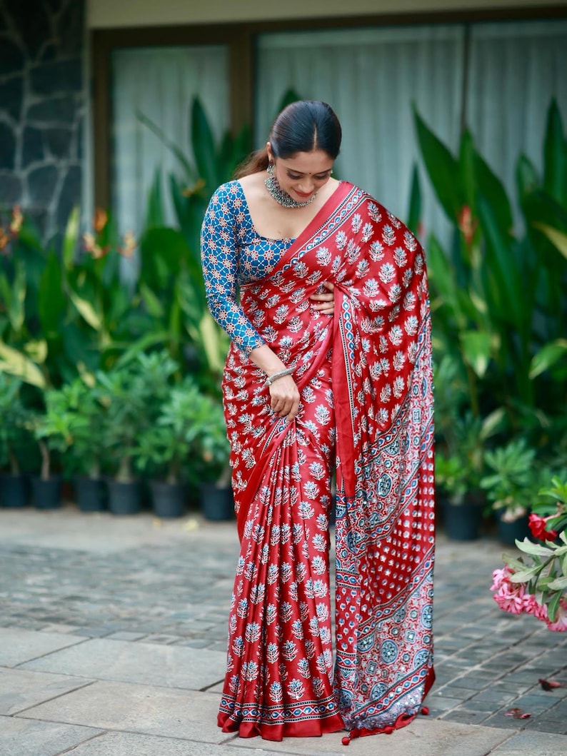 May include: A red saree featuring a repeating white floral block print pattern with a blue and white geometric patterned blouse. The saree has a wide border with intricate blue, white, and red circular motifs and a silver metallic necklace.