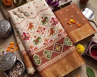 May include: A folded cream-colored saree with a diamond-patterned border featuring red, green, and orange elephant and bird motifs, paired with a rust-colored fabric piece with a gold metallic border and the text "the Queen of Silk" visible.
