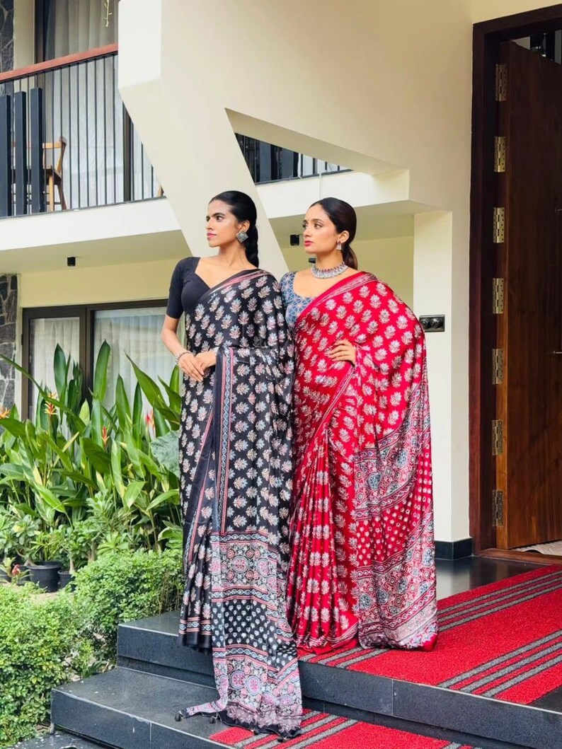 May include: Two sarees, one black with a white floral pattern and a black blouse, and one red with a white floral pattern and a blue blouse. Both garments feature intricate block-print designs, draped over the body with a long pallu end.