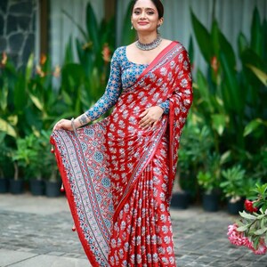 May include: A red saree featuring a repeating white and blue floral block print pattern. The garment includes a matching long-sleeved blouse with a blue and white intricate geometric design. The border displays a detailed blue and white pattern.