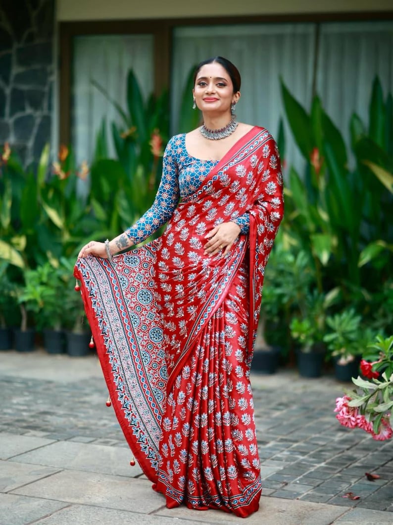 May include: A red saree featuring a repeating white and blue floral block print pattern. The garment includes a matching long-sleeved blouse with a blue and white intricate geometric design. The border displays a detailed blue and white pattern.