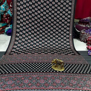 May include: A black fabric textile featuring a repeating pattern of small white and red leaf motifs, accented with a decorative border of geometric shapes and floral designs in red, white, and blue, with a small gold-colored metal cow figurine.