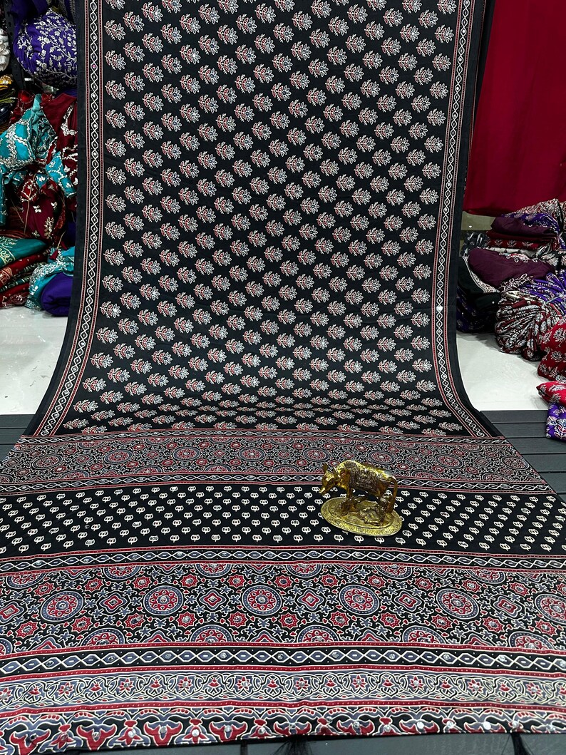 May include: A black fabric textile featuring a repeating pattern of small white and red leaf motifs, accented with a decorative border of geometric shapes and floral designs in red, white, and blue, with a small gold-colored metal cow figurine.