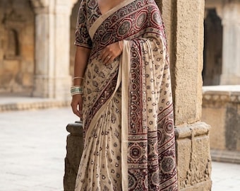 May include: A beige saree featuring a dark maroon and black block-printed pattern with circular motifs and small floral designs. The garment includes a wide decorative border with geometric shapes and a V-neck blouse with matching printed sleeves.