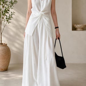 May include: A sleeveless white top featuring a V-neckline and a central front twist detail, paired with matching wide-leg trousers. The ensemble is completed by a black woven handbag and black open-toe sandals, creating a monochromatic look.