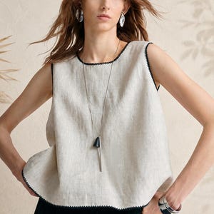 Peut inclure: Un haut sans manches en lin beige avec une encolure ronde et des emmanchures ornées de surpiqûres décoratives noires. Le vêtement est associé à un long collier argenté avec un pendentif conique noir et un large bracelet manchette argenté.