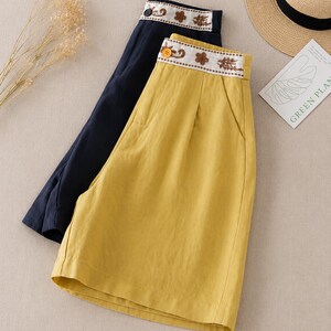 Puede incluir: Dos pantalones cortos, uno en amarillo mostaza y otro en azul marino oscuro, con dise&ntilde;o de cintura alta, una banda blanca en la cintura decorada con bordados florales marrones y un cierre de un solo bot&oacute;n en la parte delantera.