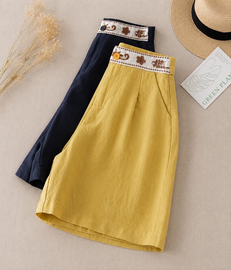 Puede incluir: Dos pantalones cortos, uno en amarillo mostaza y otro en azul marino oscuro, con dise&ntilde;o de cintura alta, una banda blanca en la cintura decorada con bordados florales marrones y un cierre de un solo bot&oacute;n en la parte delantera.