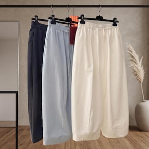 Op de afbeelding: Drie paar broeken met wijde pijpen hangen aan een zwart metalen rek. De broeken zijn uitgevoerd in de kleuren antracietgrijs, lichtblauw en off-white. Elk paar is voorzien van een elastische tailleband met een bijpassend trekkoord.