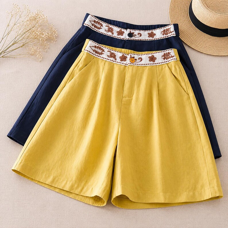 Puede incluir: Dos pares de pantalones cortos, uno amarillo y otro azul marino, con una cintura blanca con bordados florales marrones. Cada par tiene un cierre de un solo bot&oacute;n en la parte delantera central, bolsillos laterales y un dise&ntilde;o frontal plisado. Se muestran juntos.