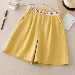Puede incluir: Pantalones cortos de color amarillo mostaza con dise&ntilde;o de cintura alta y una banda blanca decorada con bordados florales marrones. Incluyen un cierre de bot&oacute;n amarillo, bolsillos laterales y un frente plisado, ofreciendo un aspecto estructurado.