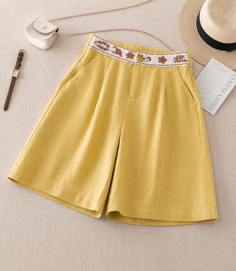 Puede incluir: Pantalones cortos de color amarillo mostaza con dise&ntilde;o de cintura alta y una banda blanca decorada con bordados florales marrones. Incluyen un cierre de bot&oacute;n amarillo, bolsillos laterales y un frente plisado, ofreciendo un aspecto estructurado.