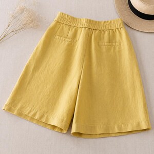 Puede incluir: Pantalones cortos de color amarillo mostaza con cintura el&aacute;stica, dos bolsillos falsos de ojal en la parte posterior y una silueta de pierna ancha. La prenda se muestra junto a un sombrero de paja con banda negra y una rama de flores secas.