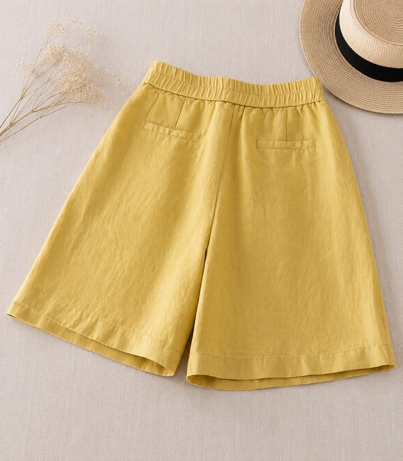 Puede incluir: Pantalones cortos de color amarillo mostaza con cintura el&aacute;stica, dos bolsillos falsos de ojal en la parte posterior y una silueta de pierna ancha. La prenda se muestra junto a un sombrero de paja con banda negra y una rama de flores secas.