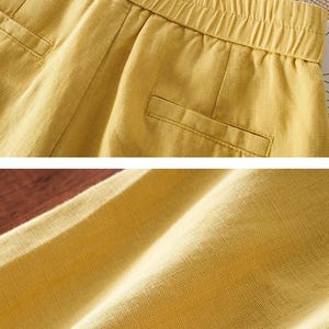 Puede incluir: Pantalones cortos de color amarillo mostaza con cintura el&aacute;stica, detalles fruncidos y dos bolsillos falsos de ojal en la parte delantera. La tela tiene una textura tejida visible y la prenda se muestra en dos vistas para resaltar la cintura y el tejido.