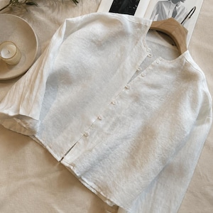 May include: A white long-sleeved button-down shirt with a round neckline, crafted from a textured fabric. The shirt features a front placket with small white buttons and long sleeves with cuffs, presented on a wooden hanger against a neutral background.