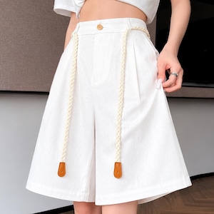 May include: White knee-length shorts featuring a high-waisted design with a single button closure, front pleats, and a decorative braided cream-colored cord belt that hangs down the front, finished with small brown leather-like tabs at the ends.