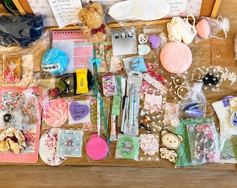 Pode incluir: Uma coleção de itens variados, incluindo um copo azul, um urso de peluche, artigos de papelaria, autocolantes, acessórios de cabelo, cartões de agradecimento, esponjas cosméticas e pequenos sacos de presente numa superfície de madeira.