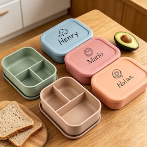 Può includere: Tre portapranzo rettangolari con coperchi in azzurro, rosa e pesca. Il coperchio blu presenta un elefante e il nome Henry, quello rosa un sole e Mario, quello pesca una ciambella e Nolan. Due contenitori aperti in verde salvia e beige.