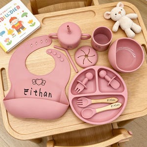 Può includere: Un set per l'alimentazione del bambino rosa, composto da bavaglino, tazza con beccuccio, ciotola, piatto e posate. Il bavaglino riporta il nome "Ethan". Sono presenti anche un piccolo orsacchiotto e un libro.