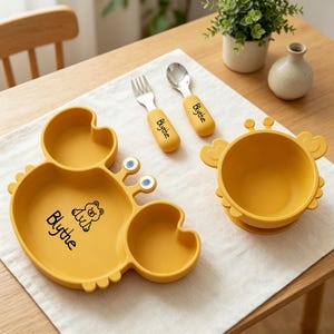 Può includere: Un set per l'alimentazione del bambino a forma di granchio giallo, con piatto, ciotola, forchetta e cucchiaio. Il piatto ha tre sezioni e una grafica di orso. Il nome "Blythe" è stampato sulle posate e sul piatto.