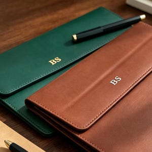 Peut inclure: Un porte-documents en cuir vert foncé et un porte-documents en cuir havane, chacun doté d'un rabat et des initiales BS dorées. Un stylo plume noir repose sur le modèle vert, et un second stylo est visible sur un bloc-notes beige à proximité.