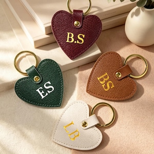 Puede incluir: Cuatro llaveros en forma de corazón con superficies texturizadas. Uno es burdeos con texto dorado B.S, uno verde oscuro con texto blanco ES, uno marrón claro con texto dorado BS y uno crema con texto dorado LB. Cada uno tiene una anilla metálica dorada.