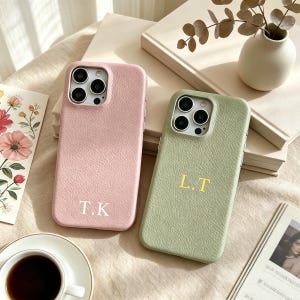May include: Two personalised phone cases. One is pale pink with the initials "T.K." in white, and the other is light green with the initials "L.T." in gold. Both cases have a textured leather-like appearance. They are on a beige surface.