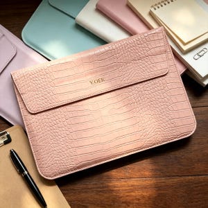 Puede incluir: Una funda para portátil de color rosa claro con un patrón texturizado de piel de cocodrilo y cierre de solapa. El texto Y.OER está grabado en letras doradas en la solapa frontal. La funda es rectangular con esquinas redondeadas.