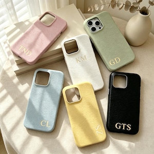 Puede incluir: Una colección de fundas personalizadas para teléfonos en varios colores, incluyendo rosa, blanco, verde, azul, amarillo y negro. Cada funda presenta una superficie texturizada e iniciales doradas. Las fundas están dispuestas sobre una superficie de tonos neutros.