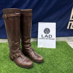 May include: A pair of knee-high brown leather boots featuring two adjustable straps with gold-toned buckles at the ankle and calf. A white sign stands behind the boots displaying a circular tree logo and the text LAD Leather Art Design.