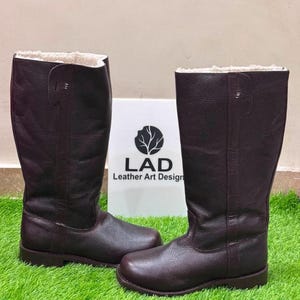 May include: A pair of dark brown, mid-calf leather boots with a textured finish and a white faux-fur lining. The boots feature a rounded toe and a flat sole. A white sign behind the boots displays a black tree logo and the text LAD Leather Art Design.