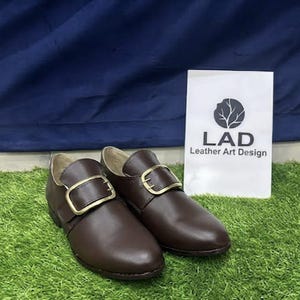 May include: A pair of dark brown leather shoes featuring a rounded toe and a prominent gold-toned metal buckle across the top. A white rectangular sign stands behind the shoes, displaying a circular tree logo and the text LAD Leather Art Design.