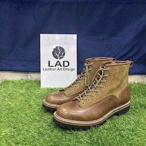 May include: A pair of brown leather and suede lace-up boots with a thick black lug sole and white stitching. A white sign behind the boots displays the text LAD Leather Art Design with a circular logo featuring a tree silhouette graphic.