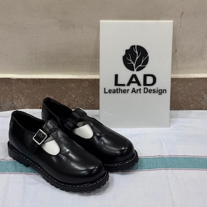 May include: A pair of black leather T-strap shoes with a silver-toned buckle and a thick lug sole. Behind the shoes stands a white rectangular sign featuring a black circular logo and the text "LAD Leather Art Design" in black lettering.