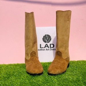 May include: Two tall, tan leather boots with a suede-like texture. The boots feature a two-tone design, with a lighter shade on the upper shaft and a darker shade on the foot and lower leg. The boots are displayed on a green surface.