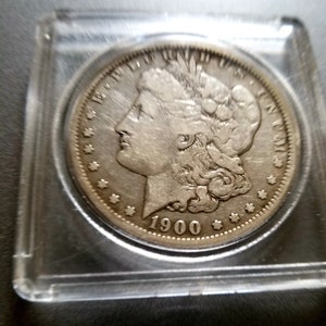 May include: A silver Morgan dollar coin with the year 1900 inscribed on the bottom. The coin features a profile of Lady Liberty with flowing hair.
