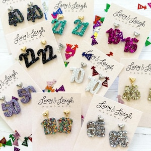 May include: Various pairs of handmade glitter earrings, each displaying a different number, such as "22", "27", "10", "50", and "65". The earrings are attached to small cards with the "Lucy & Leigh Handmade" logo, with confetti scattered around.