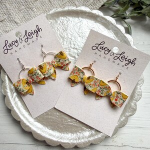 May include: Two pairs of handmade earrings. Each earring has a yellow and floral patterned bow attached to a gold hoop. The earrings are displayed on a white card with the text "Lucy & Leigh Handmade".