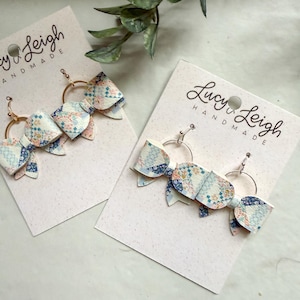Puede incluir: Dos pares de pendientes en forma de lazo con un estampado floral de patchwork en azul, rosa y blanco. Cada par está sujeto a una tarjeta de presentación blanca con la etiqueta Lucy Leigh Handmade. Cuelgan de aros metálicos con ganchos.