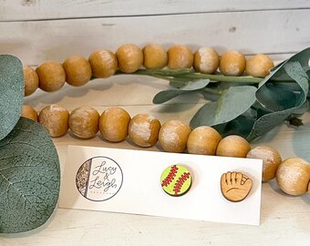 Op de afbeelding: Een paar houten oorbellen met een groene en rode softbal en een bruine honkbalhandschoen. De oorbellen liggen op een witte kaart met de tekst "Lucy & Leigh Handmade".