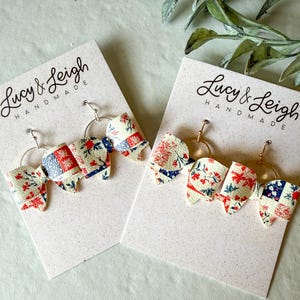 Puede incluir: Dos pares de pendientes con dijes en forma de lazo hechos de material color crema con un patrón de retazos floral rojo y azul. Cada lazo está unido a un aro de metal dorado. Los pendientes se presentan en tarjetas blancas con el nombre Lucy & Leigh.