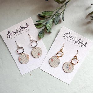 Könnte beinhalten: Zwei Paar Hängeohrringe auf weißen Präsentationskarten mit der Aufschrift Lucy & Leigh Handmade. Jeder Ohrring besteht aus einem silber- oder goldfarbenen Sechseck-Glied, das mit einer beigen, karierten Scheibe mit Blumenmuster verbunden ist.