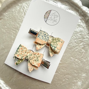May include: Two floral patterned hair bows on silver metal clips. The bows feature a mix of peach, green, and blue floral designs. The bows are attached to a white card with the text "Lucy & Leigh" in a circular logo.