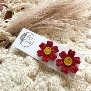 May include: Two red flower-shaped earrings with yellow centers rest on a white card. The earrings have a textured appearance. The card has the text "Lucy & Leigh" and a logo. The background is a cream-colored, textured fabric.